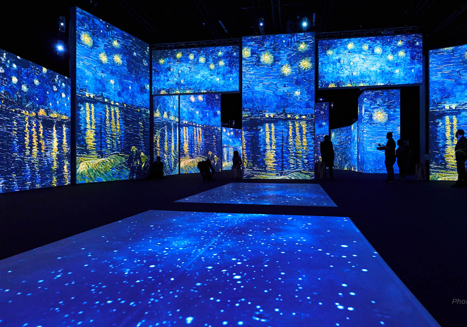 A dark room with multiple projections of Starry Nights by Vincent Van Gogh on the walls and floor. There are blue night skies with swirly stars