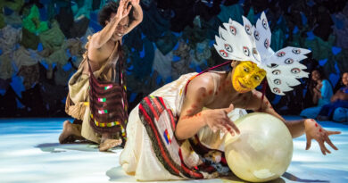 Indonesian Dancer Rianto performs in a stylised costume representing a bird with bright yellow face paint and protects a large egg. In the background the "The man who saves the world" can be seen with a bag slung across his chest and arms above head.
