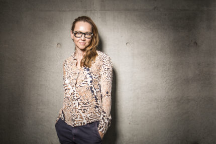 Danielle (with blonde hair and glasses) stands with her hands in her pockets in front of a concrete wall wearing a pink and black top with a collage of leopard print patterns on it and navy trousers