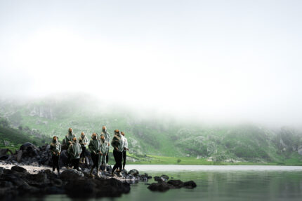 A group of children stand on the edge of the rocky shore of a lake, wearing grey hoodies, surrounded by fog. The bottom of a green mountainside is just visible in the background beneath the fog.